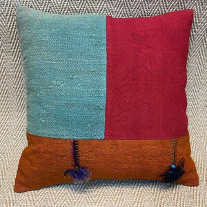 May include: A decorative pillow with a patchwork design featuring teal, red, and orange fabric. The pillow has a square shape and is accented with blue and purple tassels.