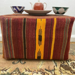 May include: A rectangular ottoman with a woven, striped design in shades of red, yellow, and brown. The ottoman is topped with a small teapot, a plate, and two decorative bowls. The bowls have blue and green patterns.
