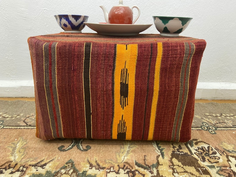 May include: A rectangular ottoman with a woven, striped design in shades of red, yellow, and brown. The ottoman is topped with a small teapot, a plate, and two decorative bowls. The bowls have blue and green patterns.