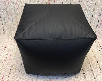 May include: A black square ottoman with stitched detailing. The ottoman is made of a smooth, leather-like material and features a cross-stitch design on the top. The cube-shaped furniture piece is suitable for a living room or bedroom.