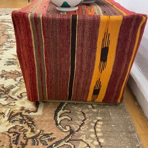 May include: A rectangular ottoman with a woven fabric cover. The fabric features vertical stripes in shades of red, orange, brown, and yellow. The ottoman is placed on a patterned rug, with a white wall in the background.