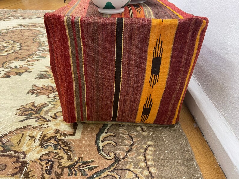 May include: A rectangular ottoman with a woven fabric cover. The fabric features vertical stripes in shades of red, orange, brown, and yellow. The ottoman is placed on a patterned rug, with a white wall in the background.