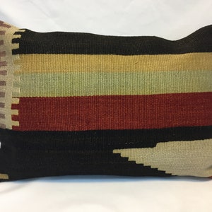 May include: Rectangular pillow with a woven design. The pillow features horizontal stripes in black, tan, gold, green, and red. The left side has a vertical pattern in tan and brown. The pillow is likely used for home decor.