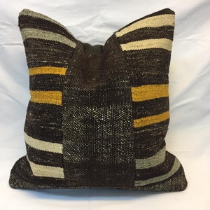 May include: Square decorative pillow with a patchwork design. The pillow features a mix of brown, yellow, and cream stripes and solid sections. The pillow is made of a woven fabric, giving it a textured appearance.