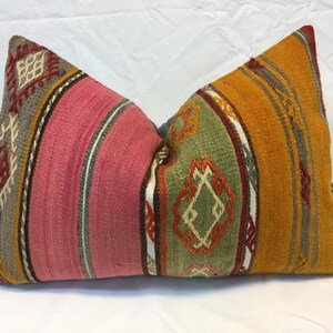 May include: A rectangular pillow with a colorful, patterned design. The pillow features a combination of pink, orange, green, and gray hues, with geometric and striped patterns. The pillow is likely made of a woven fabric.