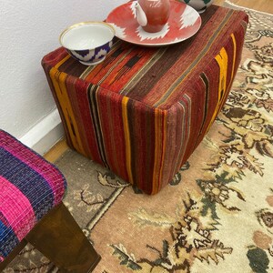 May include: A colorful, striped ottoman with a rectangular shape. The ottoman features a mix of red, orange, and brown hues. A small, patterned bowl and a red and white plate with a small teapot are on top. A second stool with pink and blue stripes is also visible.