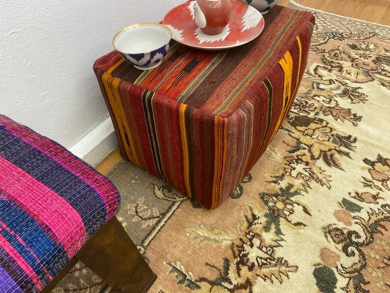 May include: A colorful, striped ottoman with a rectangular shape. The ottoman features a mix of red, orange, and brown hues. A small, patterned bowl and a red and white plate with a small teapot are on top. A second stool with pink and blue stripes is also visible.