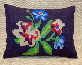 Puede incluir: Un cojín rectangular con fondo morado oscuro. Presenta un diseño floral en rojo, azul, verde y beige. Las flores tienen un diseño pixelado, lo que le da al cojín un aspecto vintage.