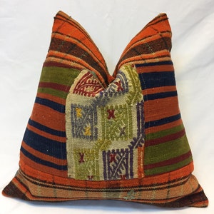 May include: A colorful patchwork pillow with a geometric pattern in shades of orange, red, green, blue, and beige. The pillow is made of woven fabric and has a fringed edge.