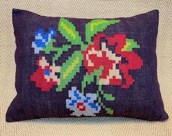 Puede incluir: Un cojín rectangular, de color púrpura oscuro, con un diseño floral colorido. Las flores bordadas presentan tonos rojos, azules, verdes y beige. El cojín es un elemento decorativo para el hogar.
