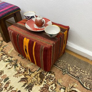 May include: A rectangular ottoman with a striped, woven fabric in shades of red, orange, and brown. A small teapot, a plate with a red and white design, and two small bowls sit on top. A bench with a colorful striped top is in the background.