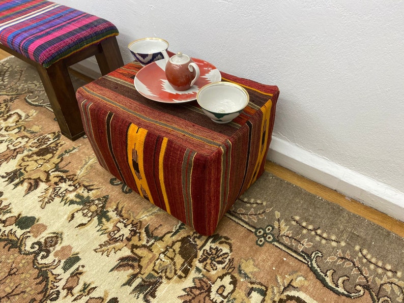 May include: A rectangular ottoman with a striped, woven fabric in shades of red, orange, and brown. A small teapot, a plate with a red and white design, and two small bowls sit on top. A bench with a colorful striped top is in the background.