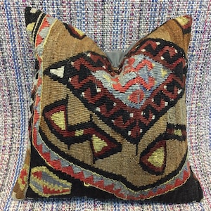 May include: A decorative pillow with a geometric pattern in shades of brown, red, black, and blue. The pillow is made of a woven fabric and has a fringed edge.