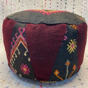May include: A round, handcrafted pouf with a flat top and a cylindrical body. The pouf features a patchwork design with burgundy, black, and patterned panels. The top has a black border with pink and yellow embroidered details. A decorative accent for any room.