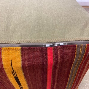 May include: A square ottoman with a striped, woven base in shades of red, yellow, and brown. The top is a solid, light green fabric, and a zipper runs along the seam. The ottoman is a decorative piece.
