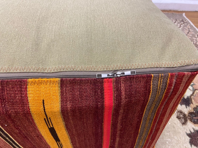 May include: A square ottoman with a striped, woven base in shades of red, yellow, and brown. The top is a solid, light green fabric, and a zipper runs along the seam. The ottoman is a decorative piece.