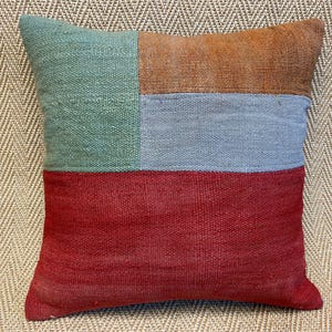 May include: A square decorative pillow with a color block design. The top section is divided into a green and orange rectangle. The middle section is a light blue rectangle, and the bottom section is red. The pillow is made of woven fabric.