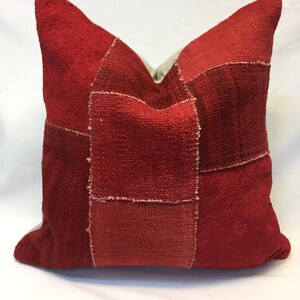 May include: A square throw pillow featuring a patchwork design composed of various shades of red textile. The surface displays a coarse, woven texture with visible seams connecting rectangular and square fabric panels in deep crimson and bright red.