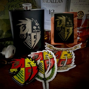 Puede incluir: Una petaca de metal negro con un escudo grabado en oro que presenta una cabeza de caballo, un árbol y el texto "Bell Au Wood". La petaca está junto a un encendedor con un diseño similar y una pegatina con un escudo similar en rojo, amarillo y verde. La pegatina tiene el texto "Darkhorse" y "Bell Au Wood".