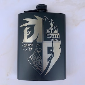 Puede incluir: Frasco de metal negro con un diseño grabado en plata de un escudo con un caballo, una amapola y las palabras "AFGHAN" e "IRAQ".