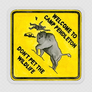 May include: A yellow warning sign with black text and a cartoon illustration. The sign reads "Welcome to Camp Pendleton, Don't Pet the Wildlife". The illustration shows a bison charging towards a person who is falling backwards.