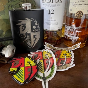 Puede incluir: Una petaca de metal negro con una tapa plateada y un diseño grabado en plata de un caballo y un escudo. El escudo tiene el texto "Bell Au Wood" y "Vie" en él. La petaca está sentada sobre una superficie de madera con tres pegatinas del mismo diseño de escudo.