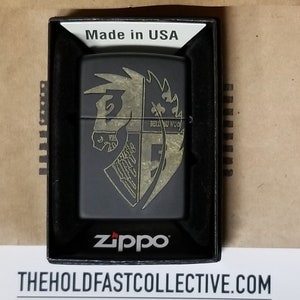 Puede incluir: Encendedor Zippo negro con un diseño dorado de una cabeza de caballo y un escudo. El encendedor está en una caja negra con el texto "Made in USA" en la parte superior. El logotipo de Zippo es visible en la parte inferior.