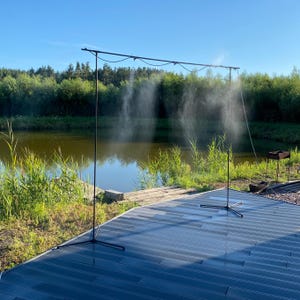 May include: A black metal frame with a misting system set up outdoors near a pond. The frame is on a grey deck with a wooden walkway leading to the water.