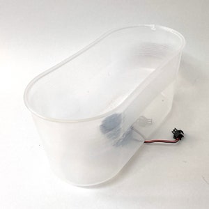 May include: A clear plastic container with a textured surface and a black wire with a connector coming out of it.