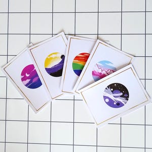 May include: Five white cards with gold trim, each featuring a different colored circle with a design inside. The circles are purple, blue, green, pink, and white with black stars.