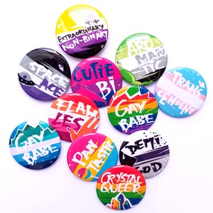 Könnte beinhalten: Zehn runde Anstecknadeln mit verschiedenen Farben und Designs. Die Nadeln zeigen Texte wie "Space Ace", "Cutie Bi", "Flawless Les", "Gay Babe", "Pan Tastic", "Demi God", "Crystal Queer", "Transcendent", "Aro Mantic" und "Extraordinary Non-Binary".