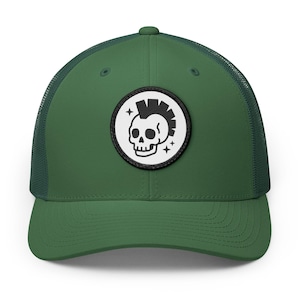 May include: A forest green trucker hat featuring a circular patch on the front panel. The patch has a white background with a black embroidered graphic of a skull with a mohawk hairstyle, accented by two small four-pointed stars on the sides.