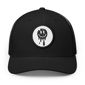 May include: A black trucker hat featuring a circular white patch on the front panel. The patch displays a black graphic of a melting smiley face with a wavy mouth. The cap has a solid front, a curved brim, and a mesh back with an adjustable strap.