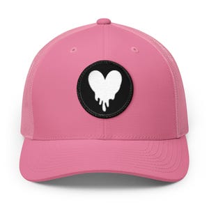 May include: A bright pink trucker cap featuring a circular black patch on the front panel. The patch displays a white embroidered heart shape with a dripping effect at the bottom. The cap has a structured front and a mesh back for ventilation.