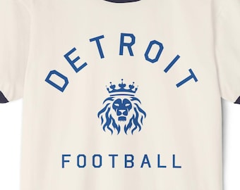 Könnte beinhalten: Ein weißes T-Shirt mit einem blauen und schwarzen Ringelkragen. Das Shirt hat ein blaues Motiv eines Löwen mit einer Krone über seinem Kopf und dem Text "DETROIT FOOTBALL" darunter.