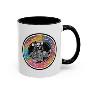 Peut inclure: Mug en céramique blanche avec intérieur et anse noirs. Le mug présente un motif circulaire avec une image en noir et blanc d'une personne jouant de la guitare et chantant, entourée du texte "QUANTUM DEPARTMENT OF TRANSPORTATION" en dégradé arc-en-ciel.