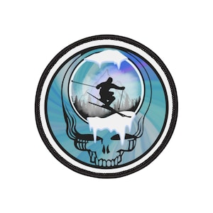 Könnte beinhalten: Runder Aufnäher mit einem stilisierten Totenkopf und einer Skifahrer-Silhouette. Das Design zeigt eine Berglandschaft und ein blau-weißes Farbschema mit schwarzem Rand.
