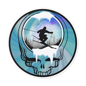 May include: Round sticker featuring a black silhouette of a skier against a mountain backdrop within a skull design. The skull has a blue and white swirl background. The skier is in mid-air, with snow and trees visible.