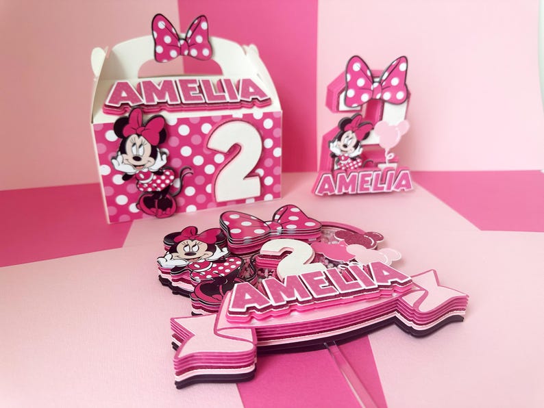 Op de afbeelding: Roze en witte taarttopper met een Minnie Mouse-figuur, het cijfer 2 en de naam Amelia. De topper is gelaagd en heeft een stippenpatroon.