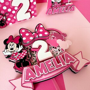 Op de afbeelding: Roze en witte gelaagde taarttopper met een Minnie Mouse-figuur, een strik, het cijfer 2 en de naam Amelia. De topper staat op een roze achtergrond.