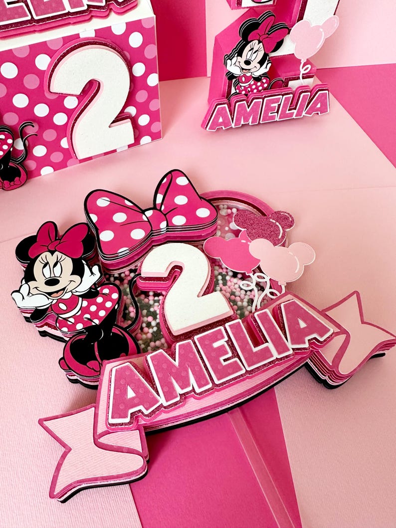 Op de afbeelding: Roze en witte gelaagde taarttopper met een Minnie Mouse-figuur, een strik, het cijfer 2 en de naam Amelia. De topper staat op een roze achtergrond.