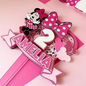 Op de afbeelding: Roze en witte taarttopper met een Minnie Mouse-figuur, een wit cijfer 2 en de naam Amelia. De topper is versierd met roze en witte stippen en glitter.
