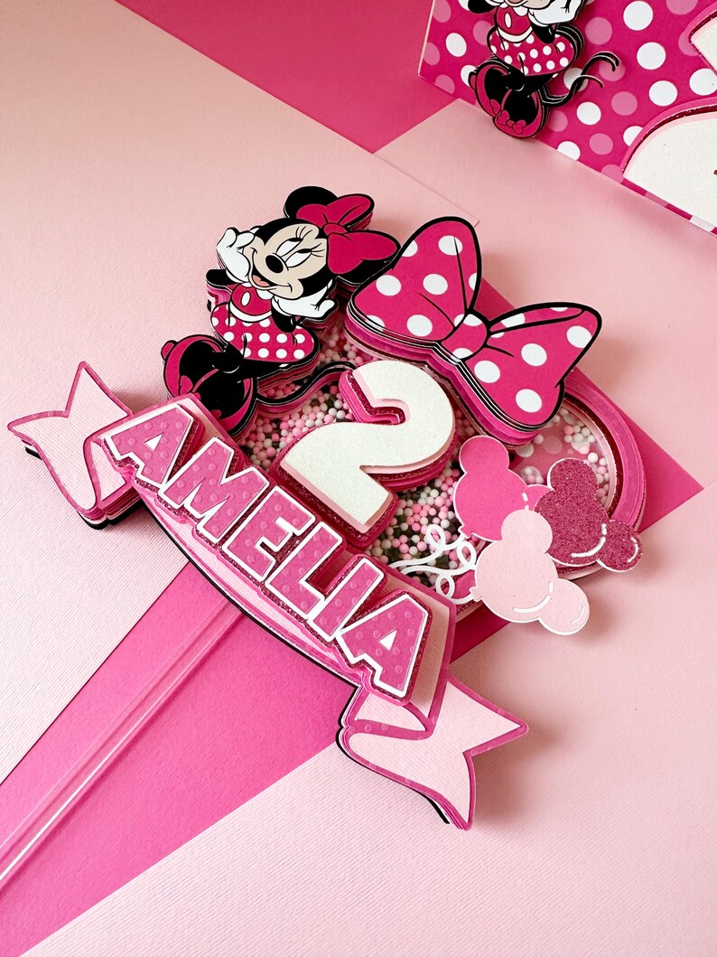 Op de afbeelding: Roze en witte taarttopper met een Minnie Mouse-figuur, een wit cijfer 2 en de naam Amelia. De topper is versierd met roze en witte stippen en glitter.