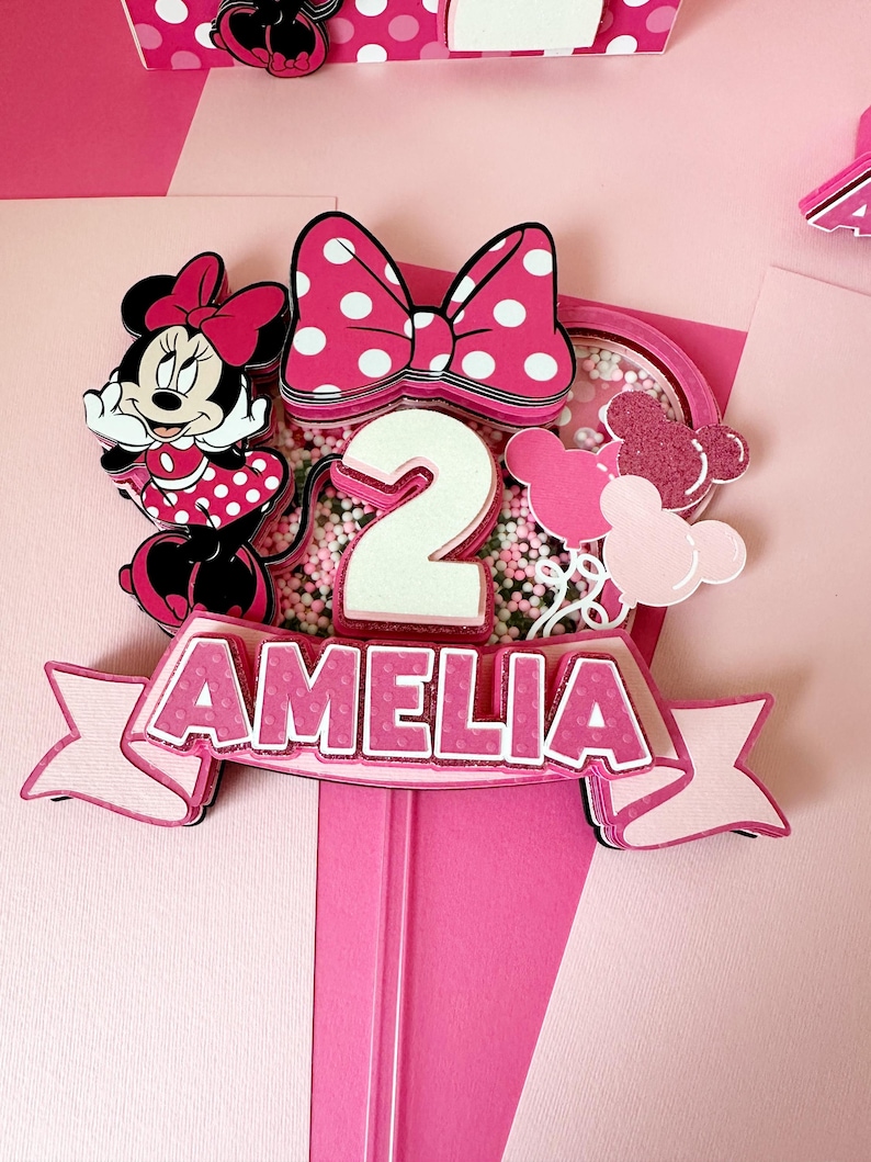 Op de afbeelding: Een roze en witte taarttopper met een cartoon Minnie Mouse, een wit cijfer 2, een roze strik met witte stippen en de naam Amelia in roze letters met witte stippen.