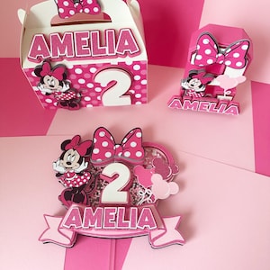 Op de afbeelding: Roze en witte Minnie Mouse-thema feestdecoraties met de naam Amelia en het nummer 2. De decoraties omvatten een cadeaubox, een taarttopper en een nummer 2 middelpunt.