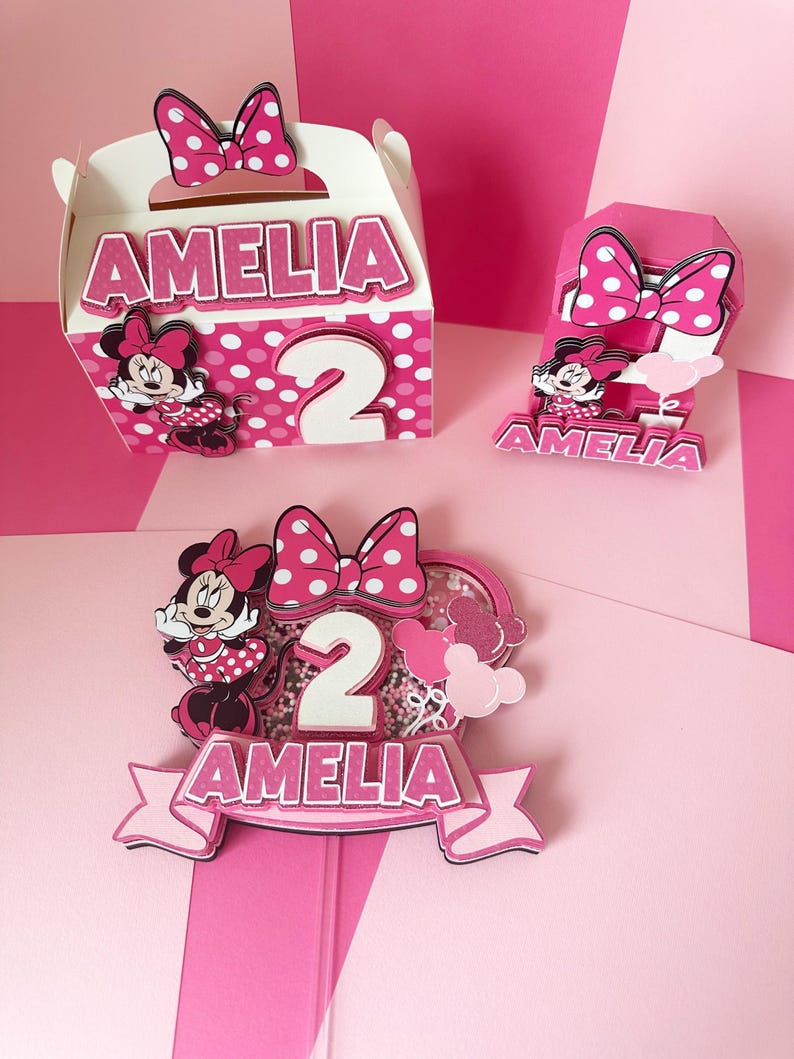 Op de afbeelding: Roze en witte Minnie Mouse-thema feestdecoraties met de naam Amelia en het nummer 2. De decoraties omvatten een cadeaubox, een taarttopper en een nummer 2 middelpunt.