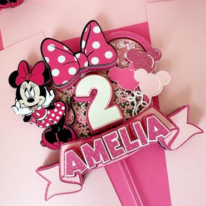 Op de afbeelding: Een roze en witte Minnie Mouse-thema caketopper met een strik, een nummer 2 en de naam Amelia. De topper is versierd met roze en witte sprinkles.