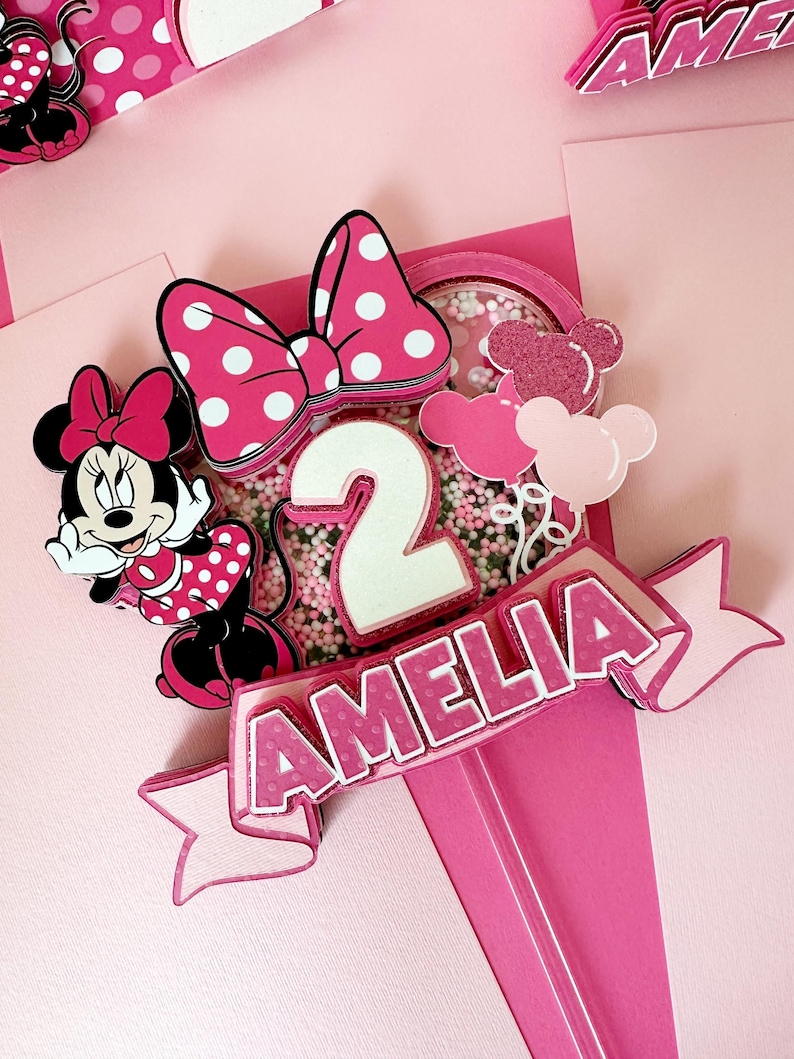 Op de afbeelding: Een roze en witte Minnie Mouse-thema caketopper met een strik, een nummer 2 en de naam Amelia. De topper is versierd met roze en witte sprinkles.