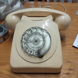 Könnte beinhalten: Ein cremefarbenes Vintage-Wählscheibentelefon mit einem oben aufliegenden Hörer. Die kreisförmige Wählscheibe verfügt über durchsichtige Kunststofflöcher, um die in der Mitte die schwarzen Ziffern null bis neun angeordnet sind.