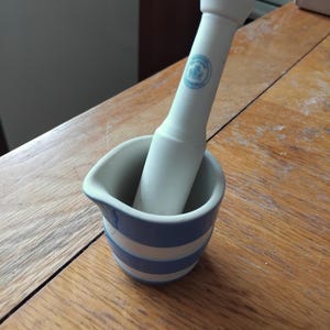 Könnte beinhalten: Ein weißes Keramik-Mörser- und Stößel-Set. Der Mörser hat blaue und weiße horizontale Streifen und einen Ausguss. Der Stößel ist weiß mit einem blauen Emblem. Das Set steht auf einer Holzoberfläche.